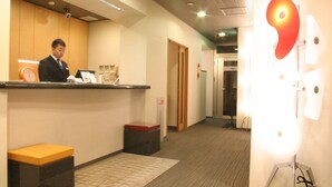 Een hotel receptie met een receptiebalie, een klok, een afvalbak en een wandlamp.