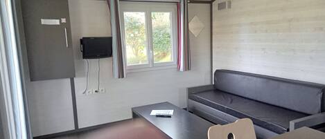 A compact room with a sofa, a small table, a television mounted on the wall, and a window with curtains.