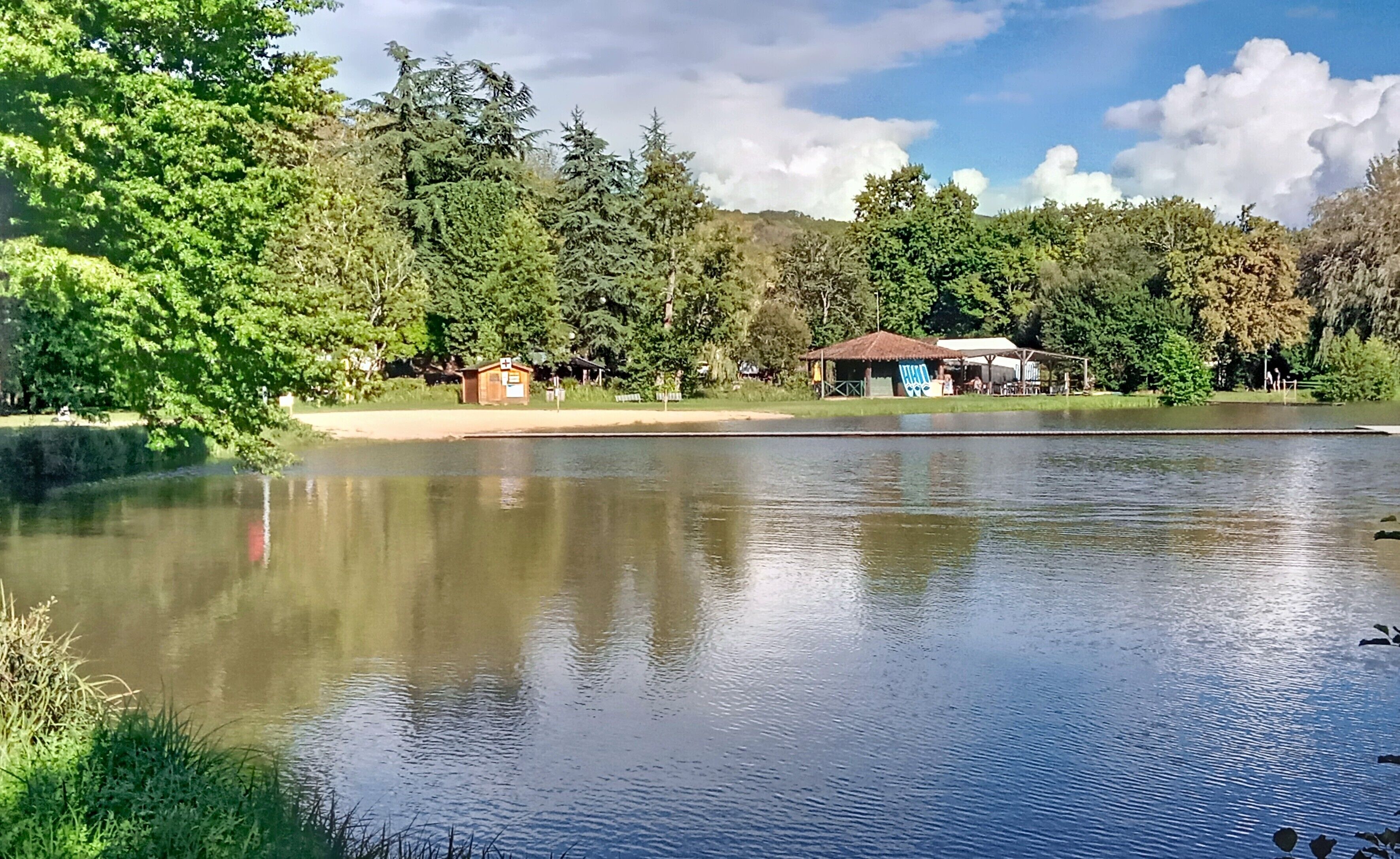 A calm lake with a small dock, surrounded by lush greenery and a few buildings in the background.