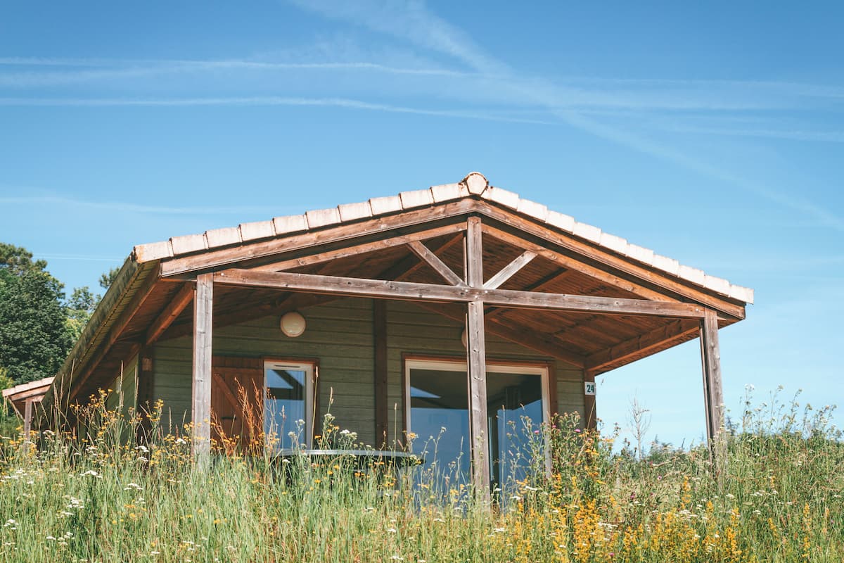 A rustic wooden cabin with a pitched roof and a porch, surrounded by tall grass and wildflowers.