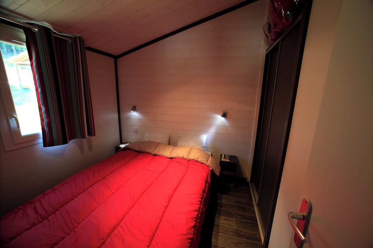 A bedroom with a red bedspread, two bedside lamps, a nightstand, and a window with curtains.