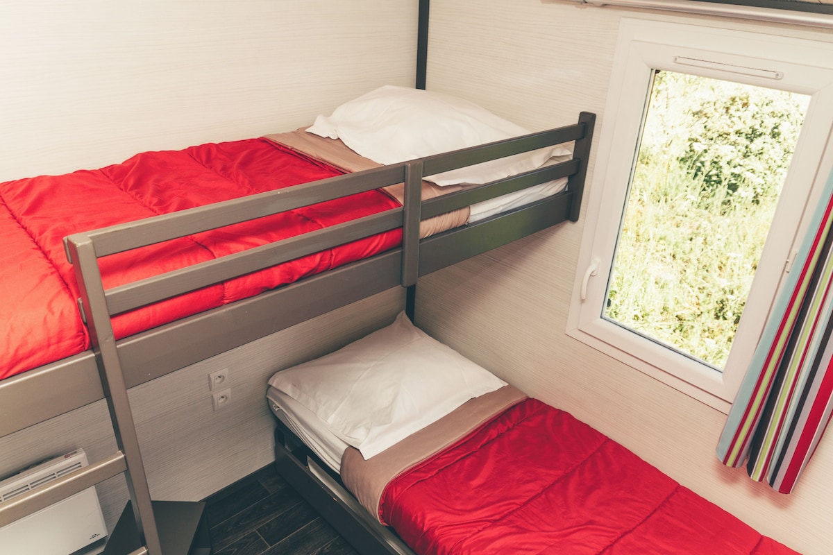 A bunk bed with red bedding, a window with a view of greenery, and a small desk with drawers.