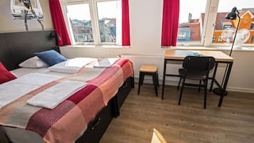 Quarto com cama, escrivaninha e cadeira. Janelas com cortinas vermelhas, radiador e luminária na parede.
