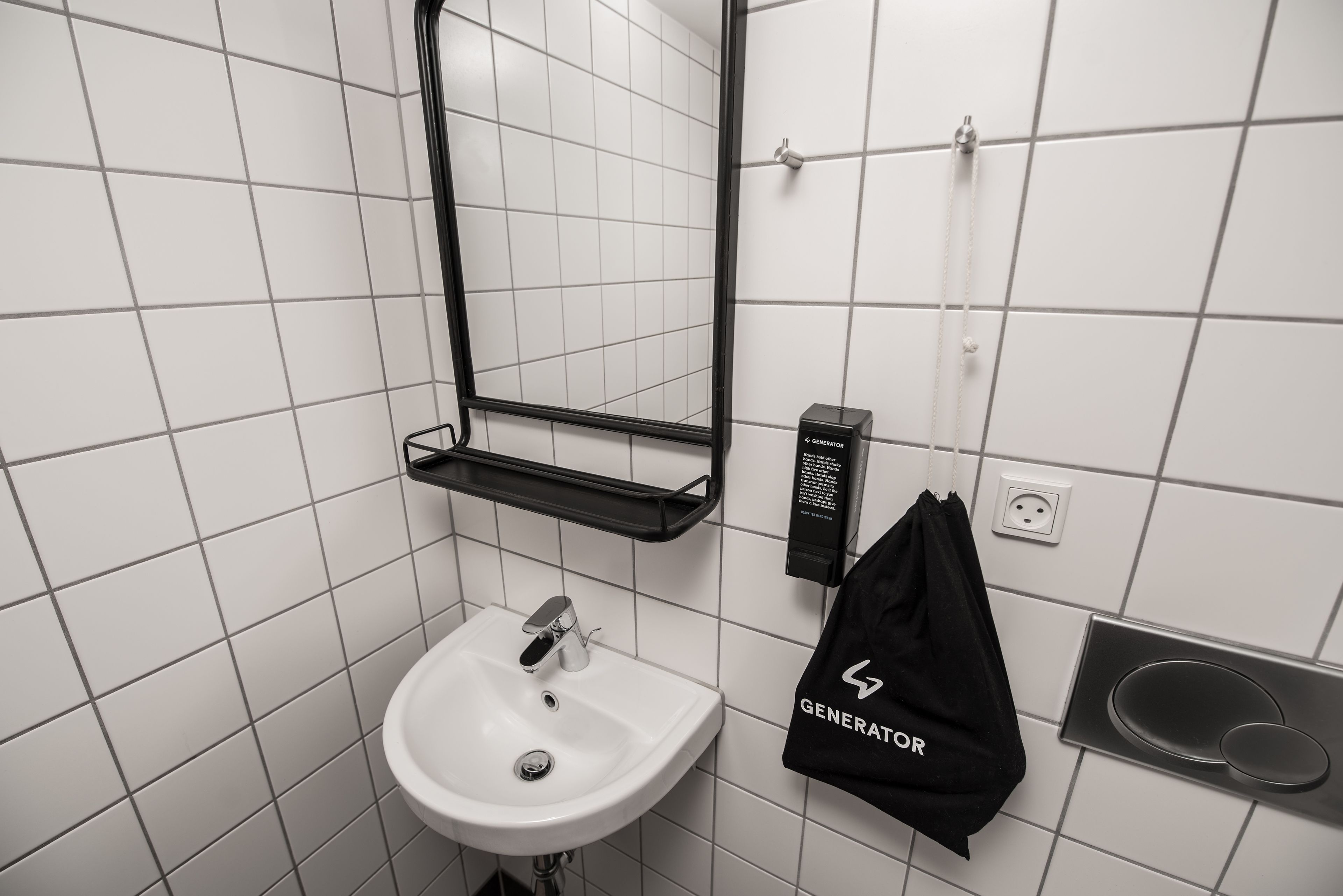 Un baño con un lavabo blanco, un espejo, un bolso negro colgando en la pared y un estante negro sobre el lavabo.