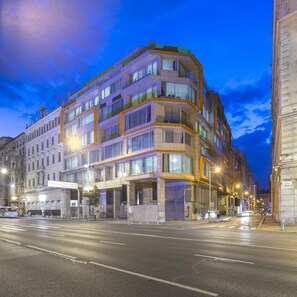Un hôtel à plusieurs étages, doté d’un design moderne, qui se distingue par son emplacement en angle, ses grandes fenêtres et sa façade bien éclairée.