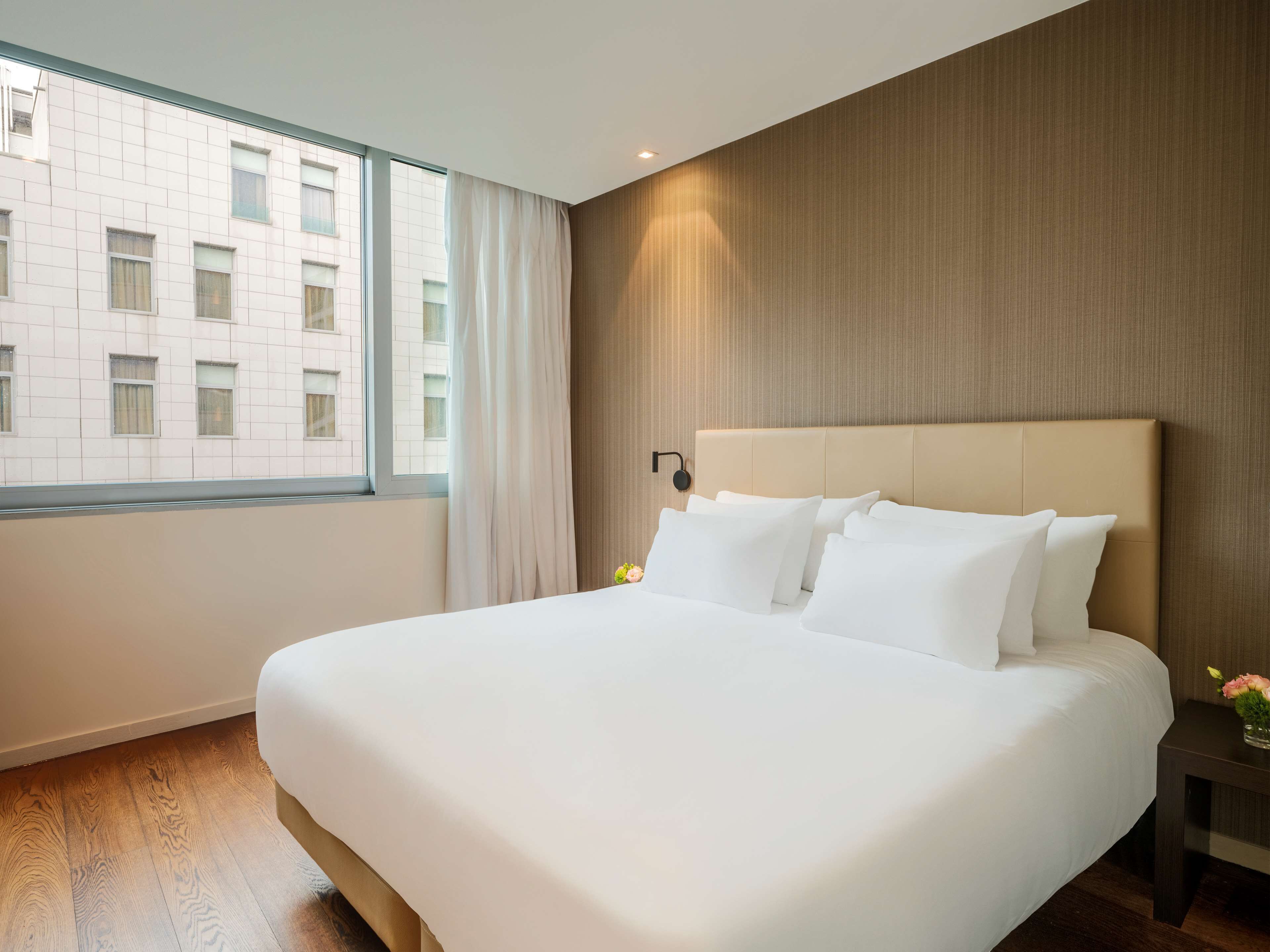 Una habitación de hotel moderna con una cama grande, una mesita de noche, una lámpara de pared y un ventanal con cortinas.