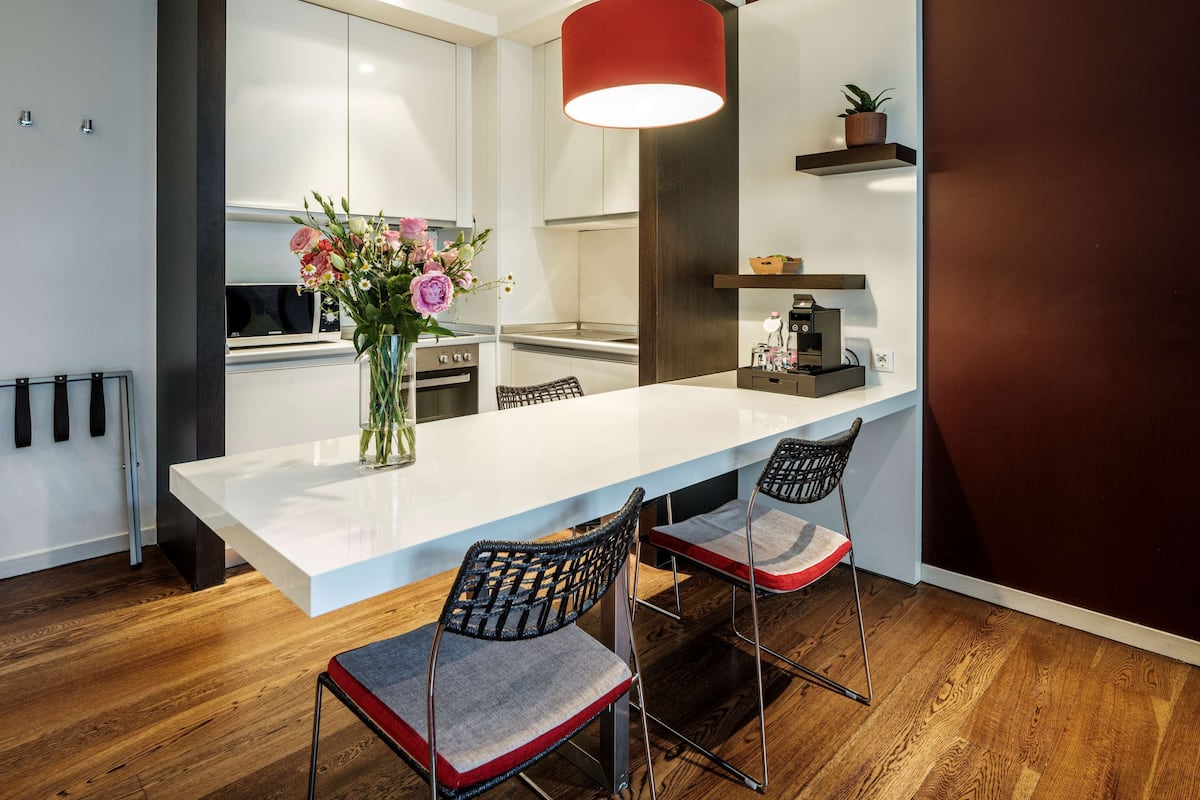A modern kitchen with a white countertop, two bar stools, a red pendant light, and a vase of flowers.