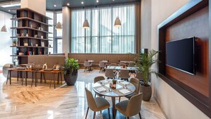 A modern dining area with a large window, a flat-screen TV, and a bookshelf. - NH Collection Budapest City Center (Budapest)
