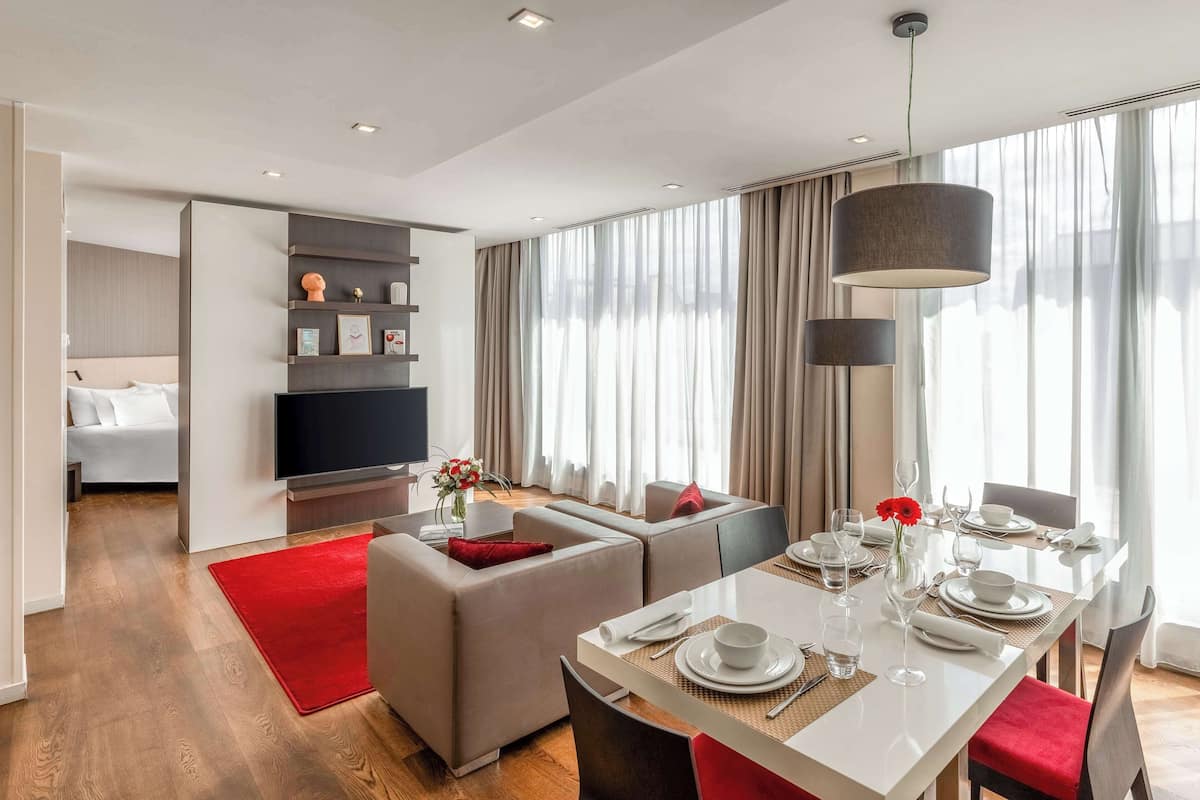 A modern hotel room with a dining area, a sofa, a TV, and a large window with curtains.