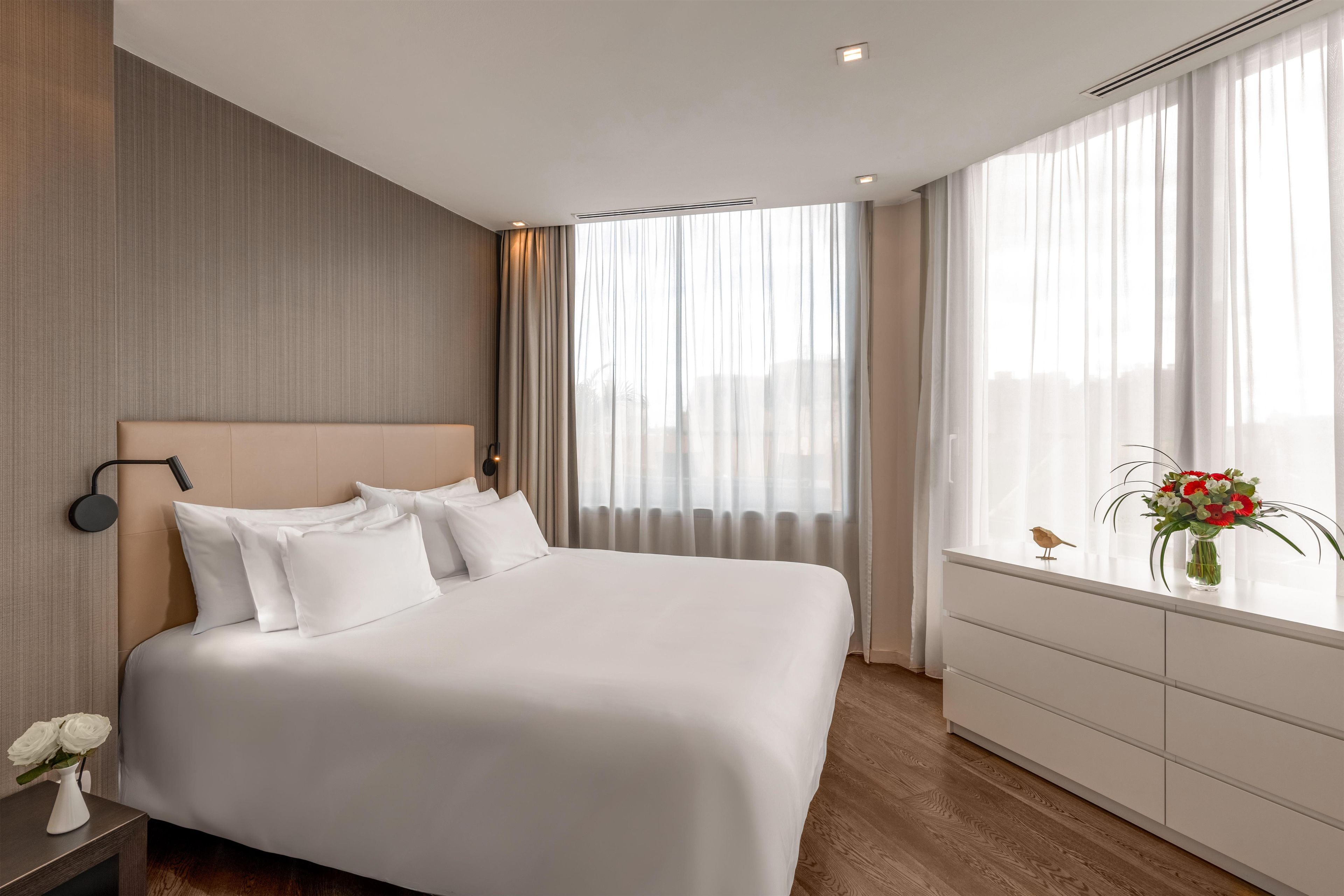 A hotel room with a large bed, a bedside table with a lamp, a dresser, and a window with curtains.