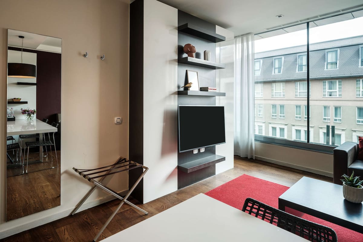A modern living room with a large window, a flat-screen TV, and a dining area with a red rug.