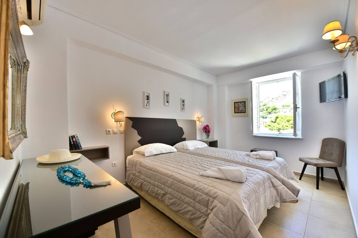 A hotel room with two beds, a desk, a chair, a TV, and a window with a view of greenery.