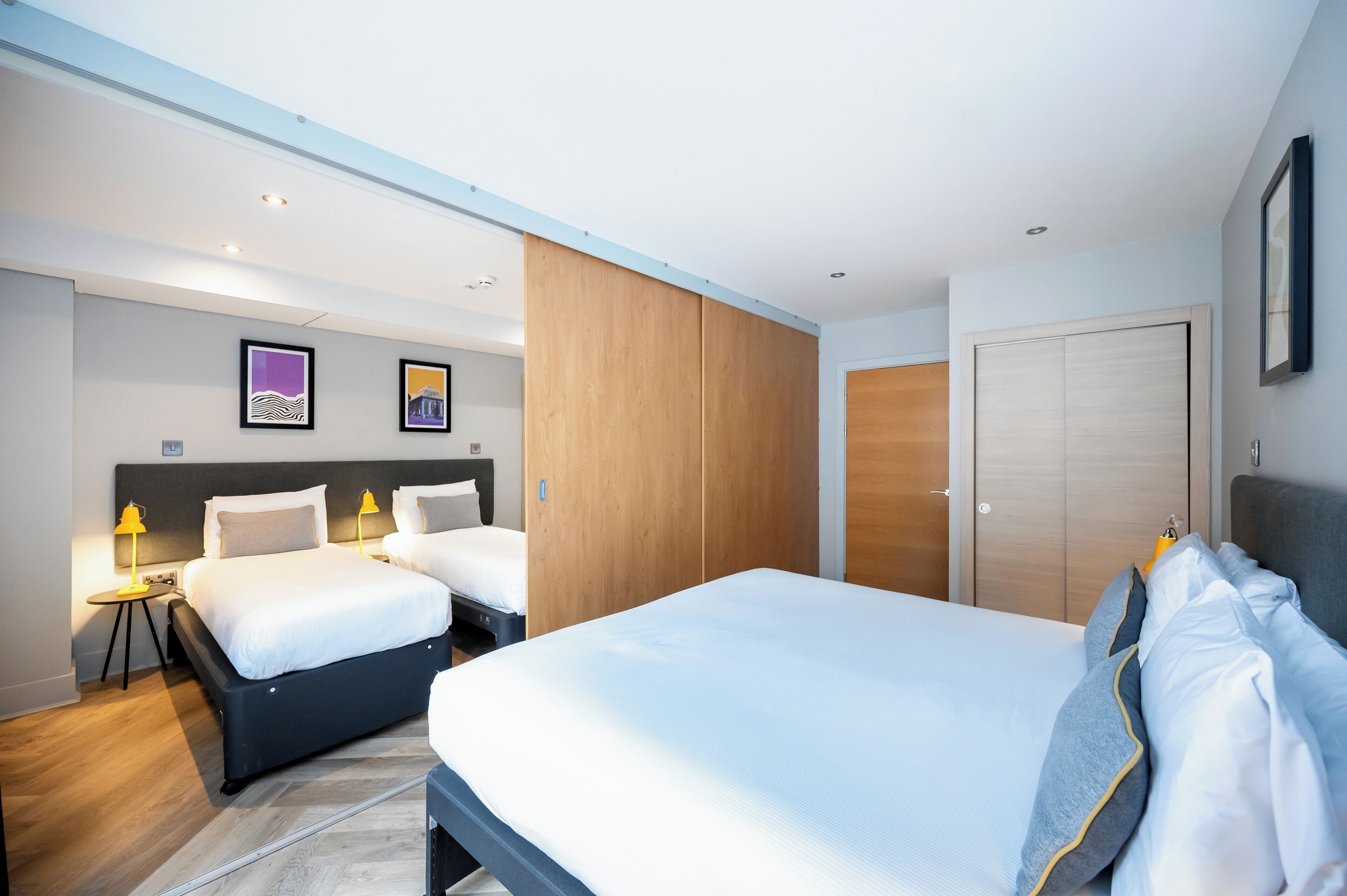 Une chambre d’hôtel moderne avec deux lits, une armoire en bois et une autre armoire avec un miroir, ainsi qu’une porte dotée d’une poignée.