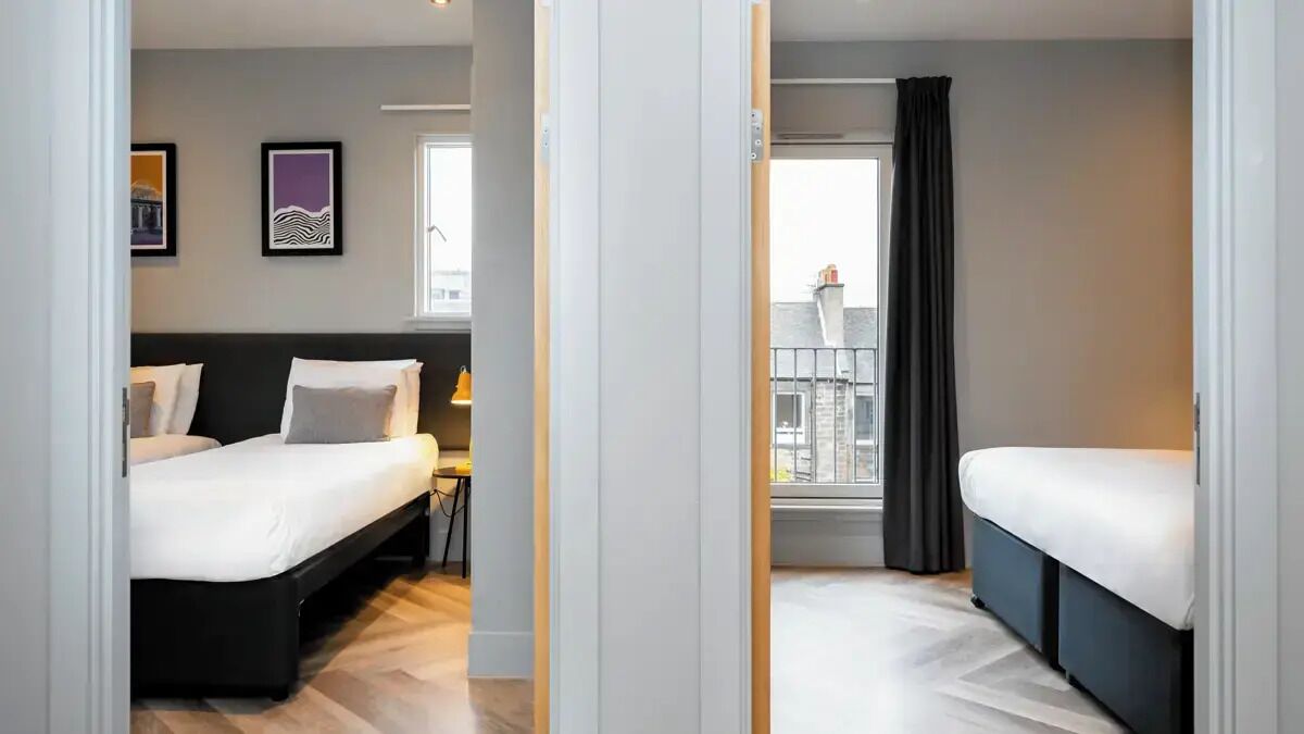 A hotel room with two beds, a desk, and a window with a view of buildings.