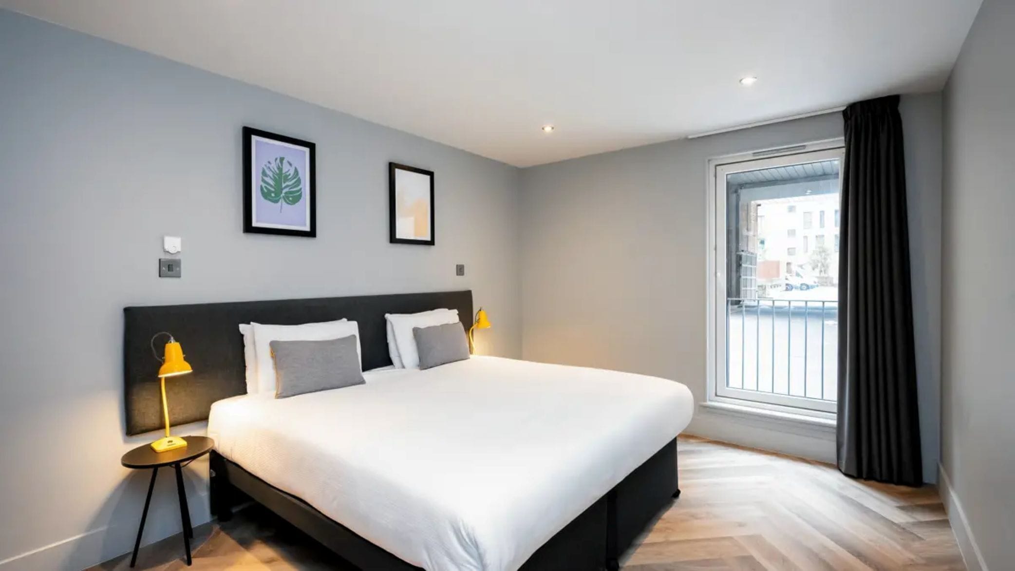 A modern hotel room with a large bed, two bedside lamps, a window with curtains, and two framed pictures on the wall.