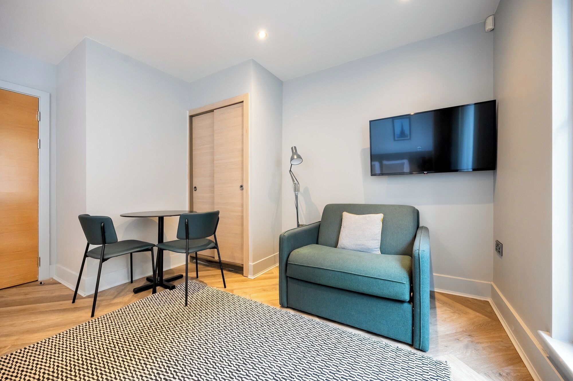 Ein modernes Wohnzimmer mit einer Couch, zwei Sesseln, einem kleinen Tisch, einer Stehlampe und einem an der Wand befestigten Fernseher.