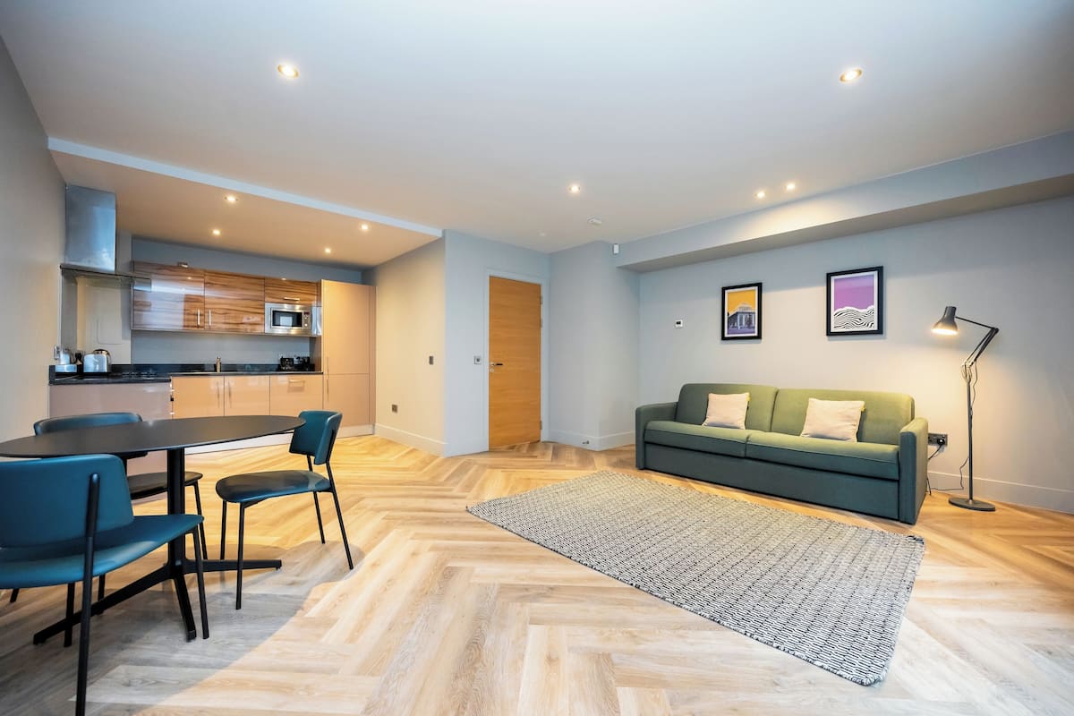 A modern living room with a green sofa, a dining table with chairs, a patterned rug, and a kitchen in the background.