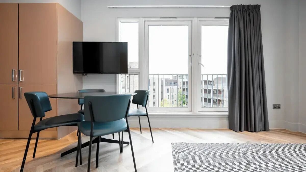 A modern living room with a flat-screen TV, a dining table with chairs, a large window with curtains, and a view of buildings.
