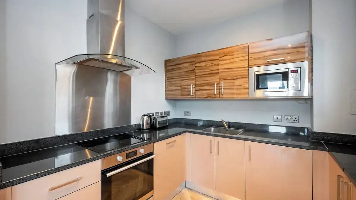 Una cocina moderna con campana de acero inoxidable, armarios de madera, microondas y encimera negra.