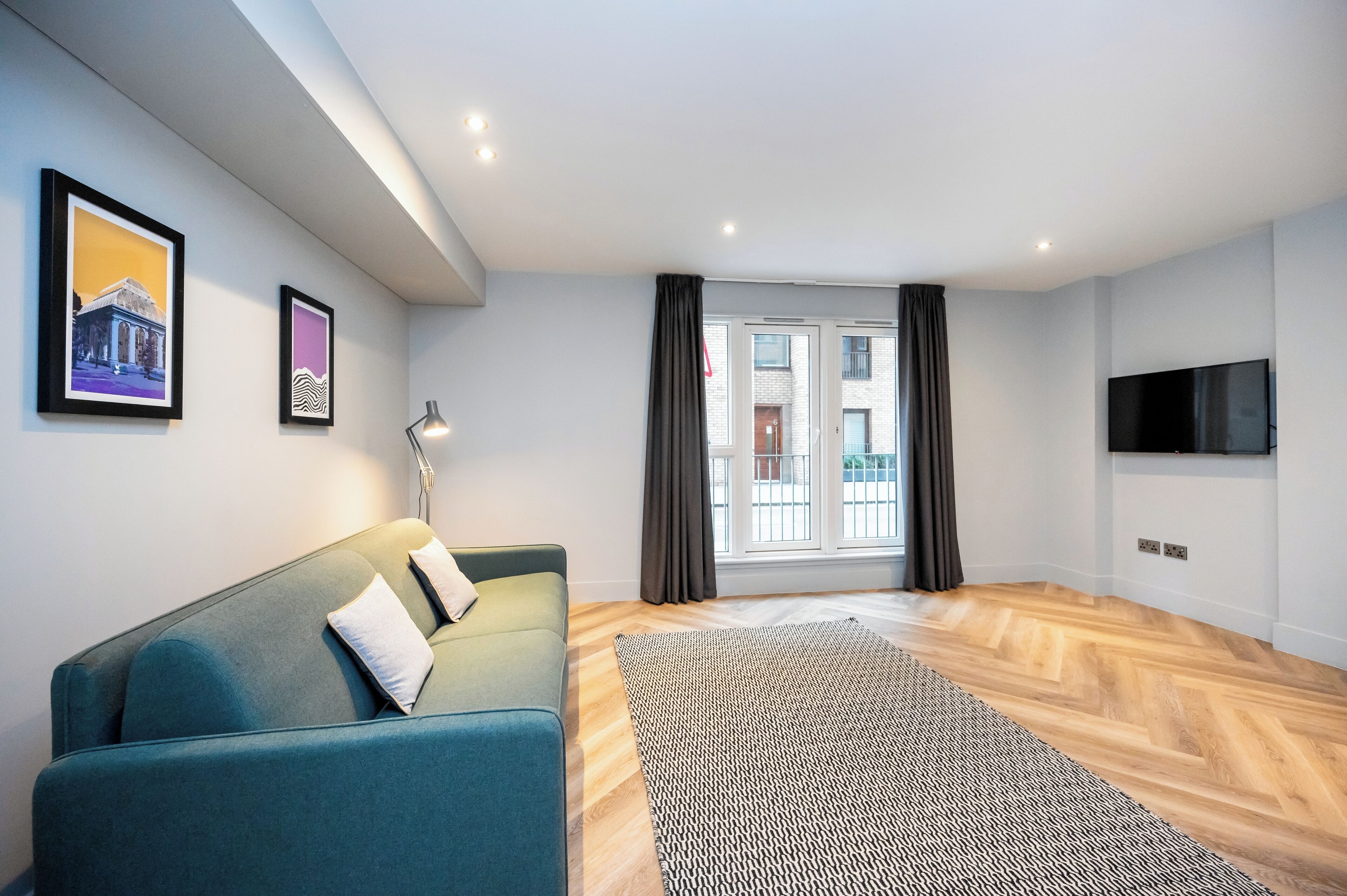 Un salon moderne avec un canapé de couleur turquoise, un tapis à motifs, un téléviseur à écran plat et des œuvres d’art encadrées accrochées au mur.