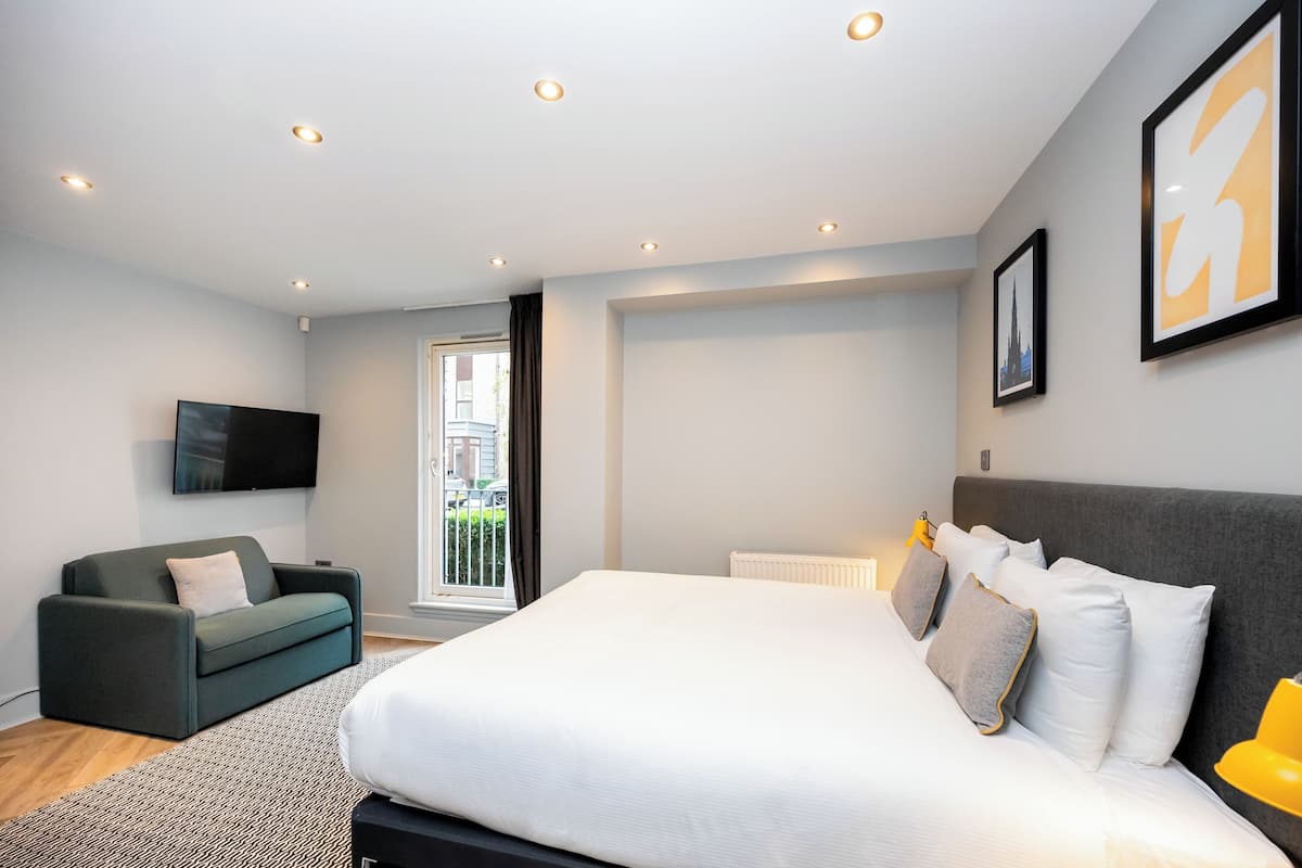 A modern bedroom with a large bed, a grey sofa, a flat-screen TV, and a window with a view of buildings.