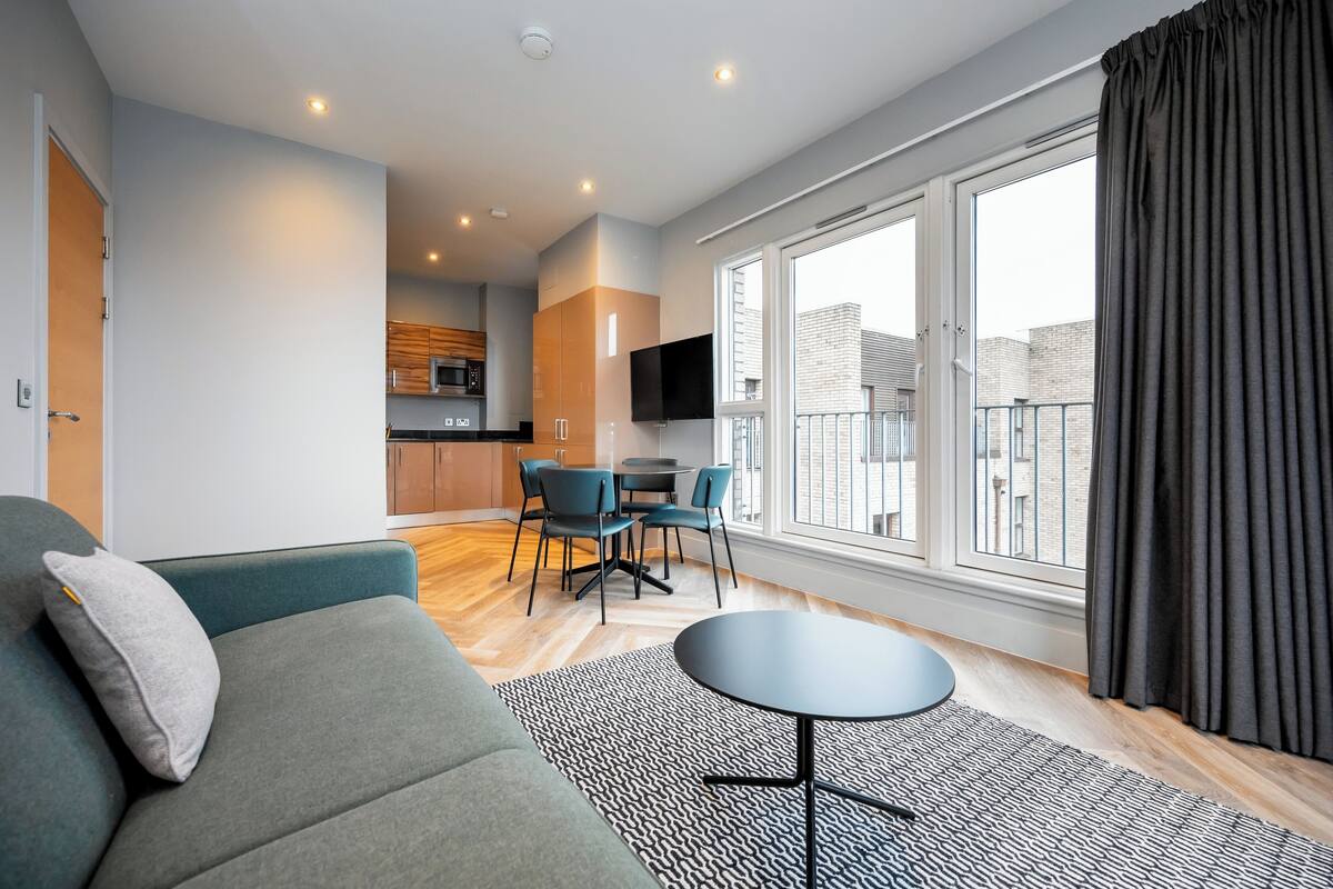A modern living room with a sofa, a coffee table, and a dining area with chairs. There is a kitchen in the background with a TV mounted above the counter.