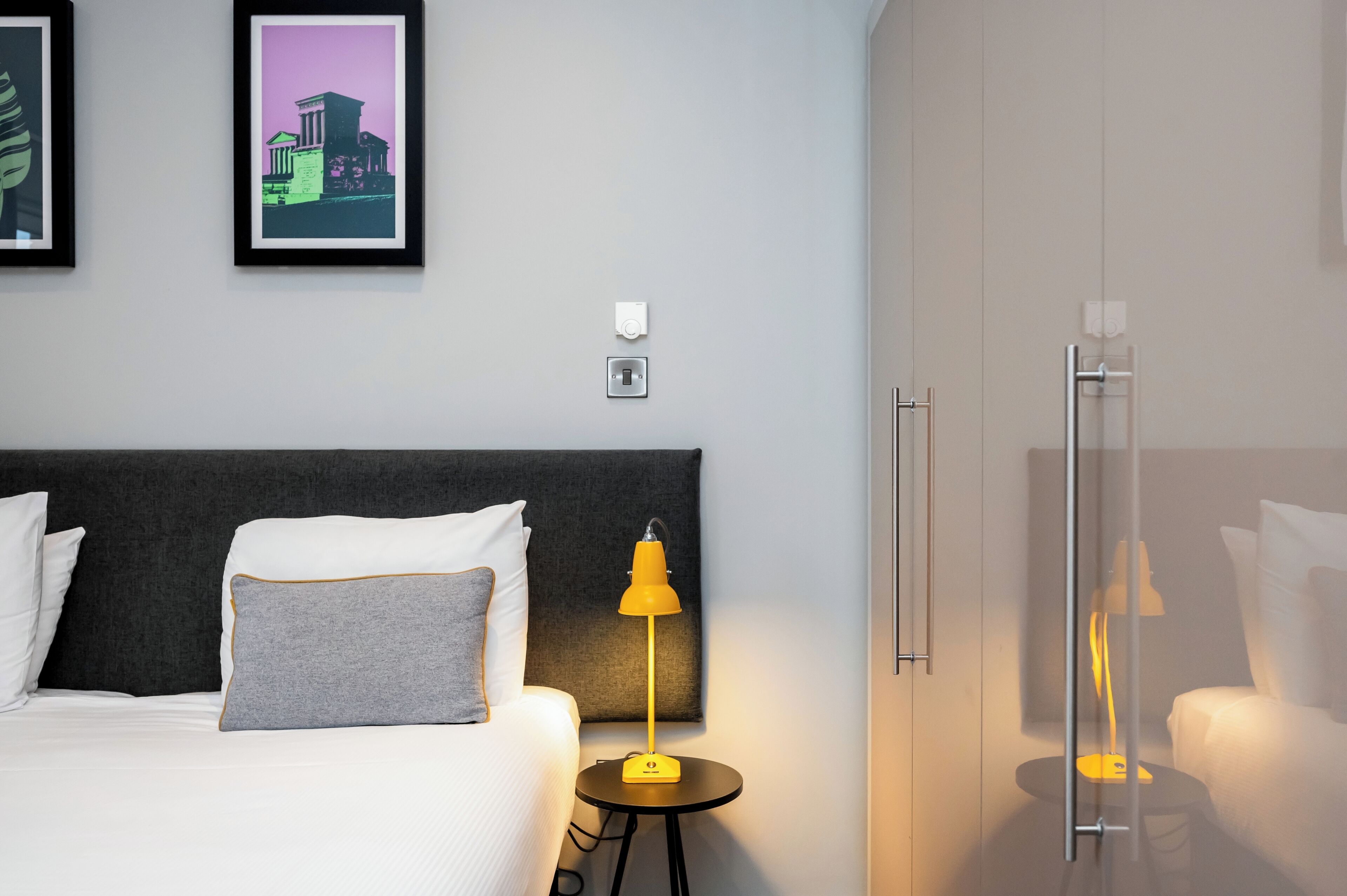 Une chambre d’hôtel avec un lit, une table de chevet avec une lampe, une armoire et deux tableaux encadrés au mur.