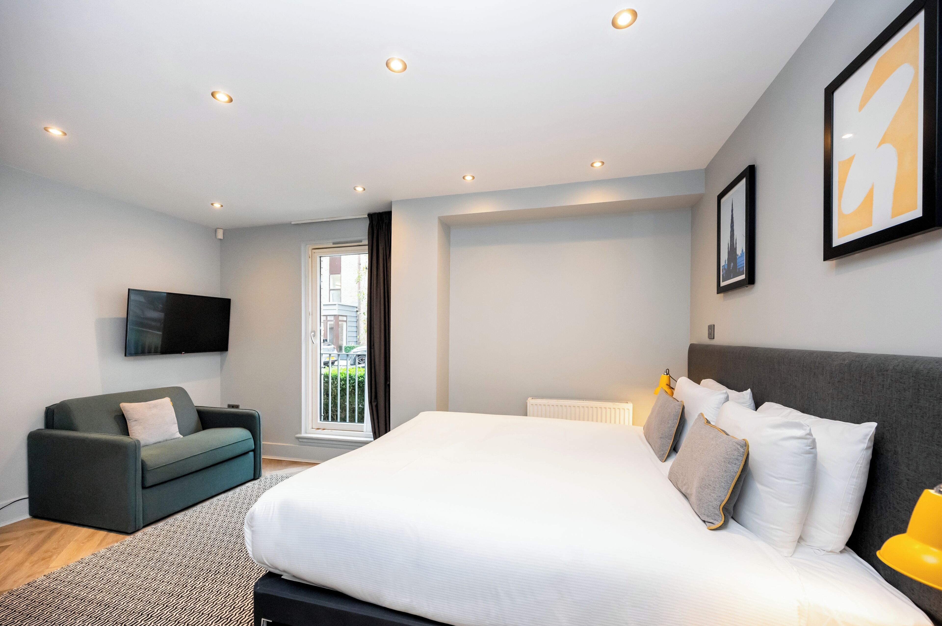 A modern bedroom with a large bed, a grey sofa, a flat-screen TV, and a window with a view of buildings.