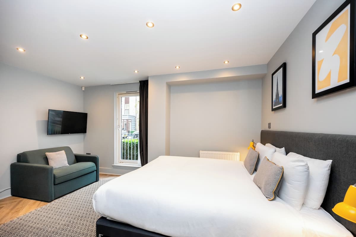 A modern bedroom with a large bed, a grey sofa, a flat-screen TV, and a window with a view of buildings.