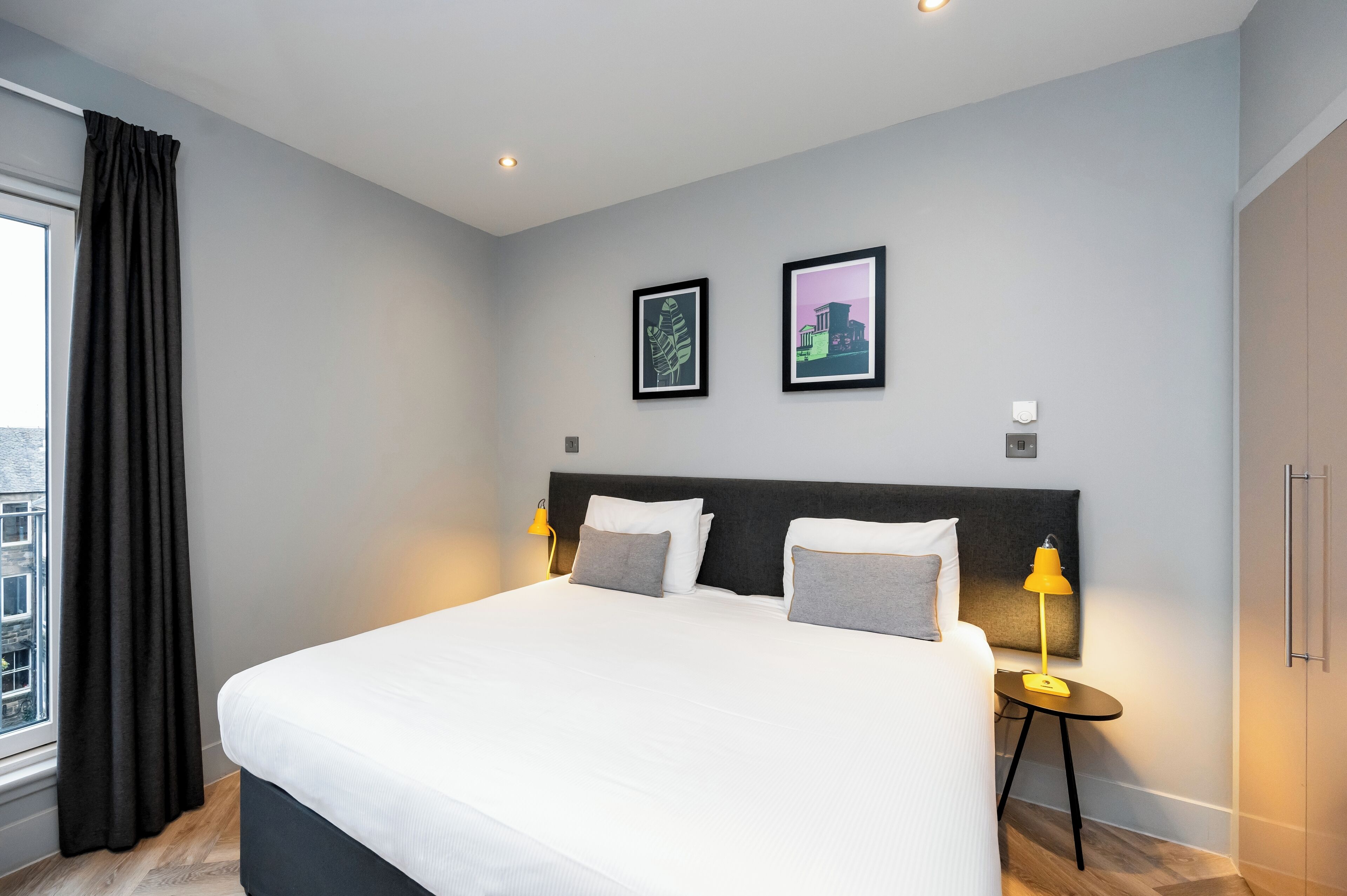 A modern hotel room with a large bed, two bedside lamps, a window with curtains, and two framed pictures on the wall.