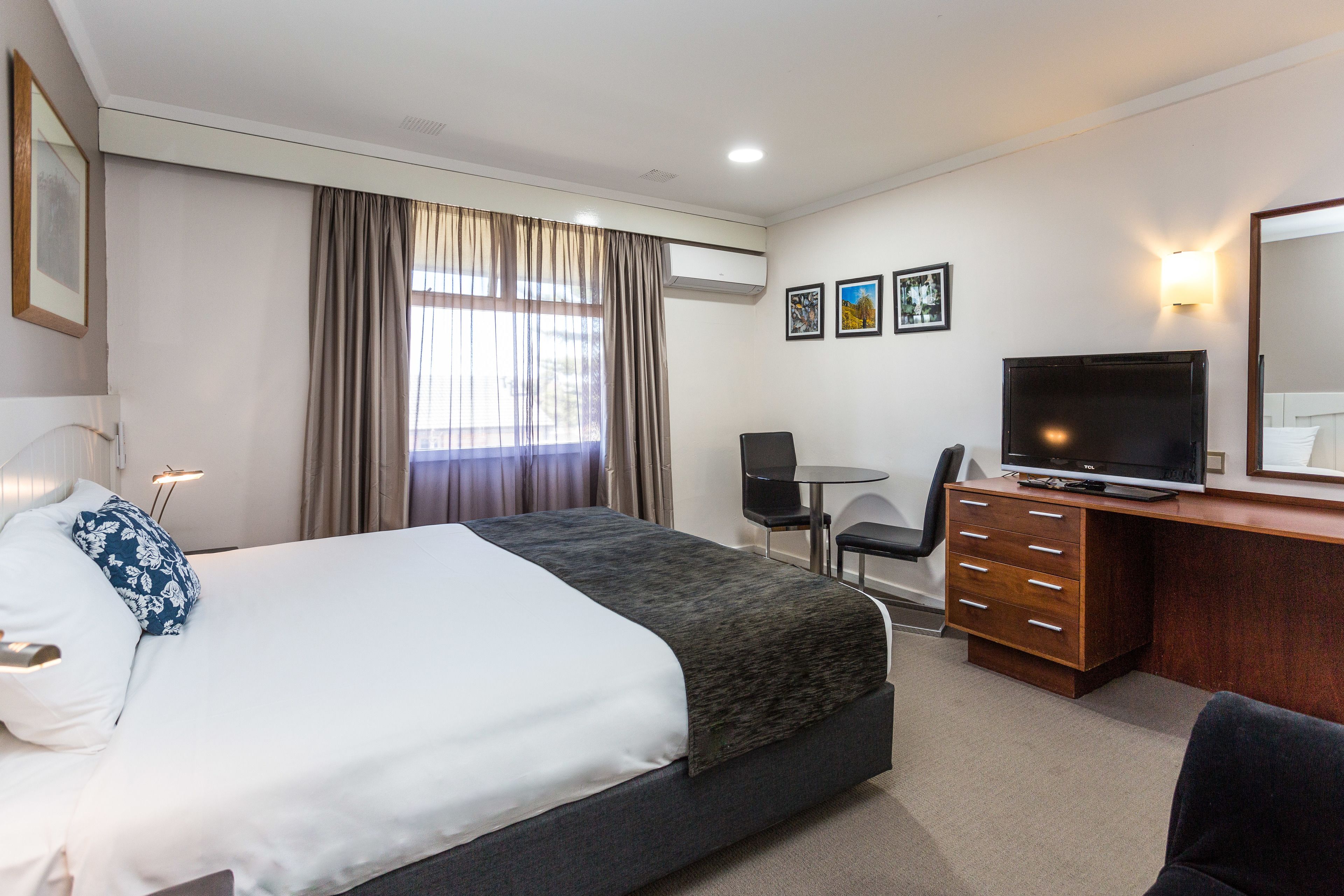 A hotel room with a bed, a desk with a television, a chair, and a desk lamp.