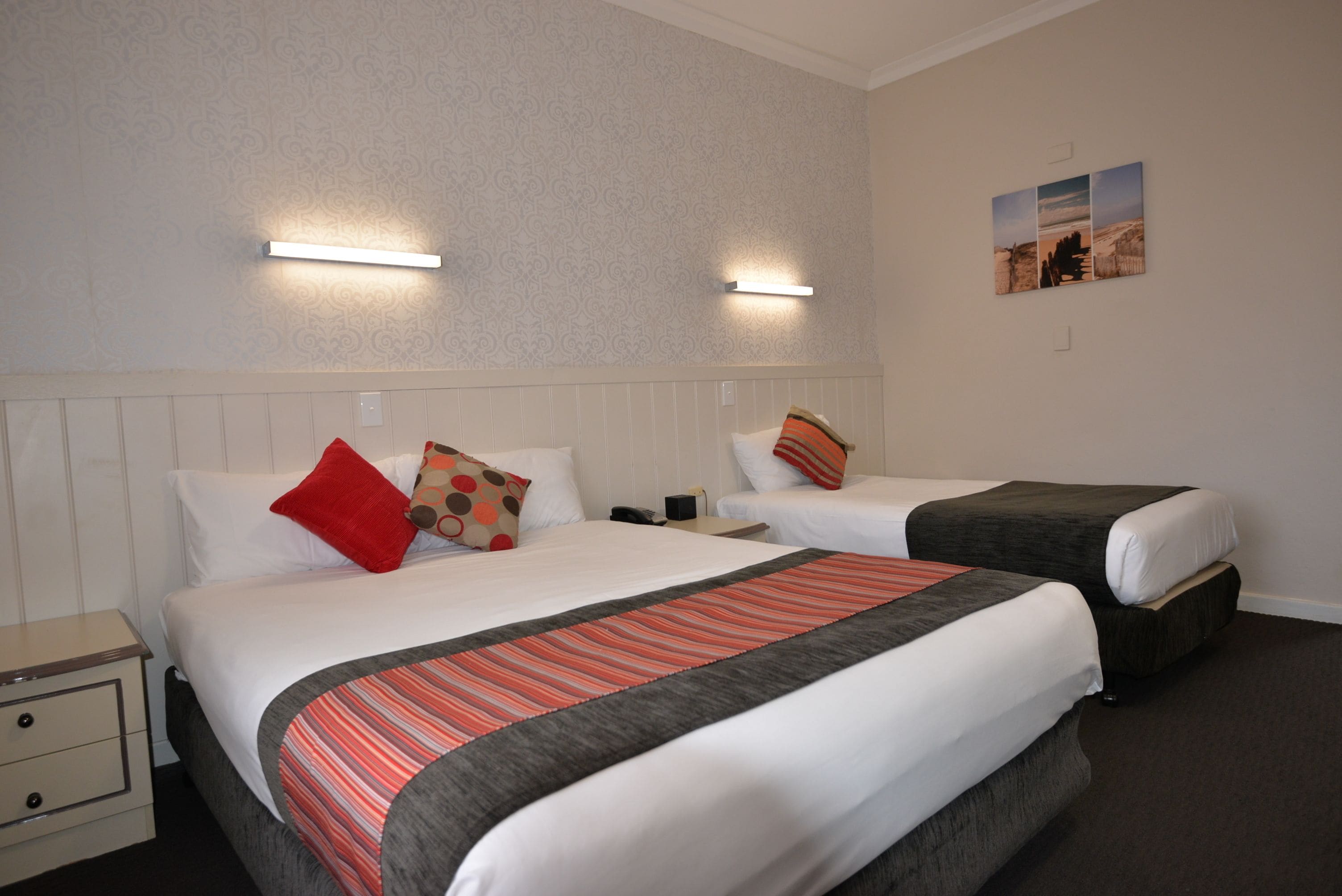 Habitación de hotel con dos camas, cada una con almohada y manta. Hay una mesita de noche con lámpara y teléfono. La habitación tiene un papel tapiz estampado y un cuadro en la pared.