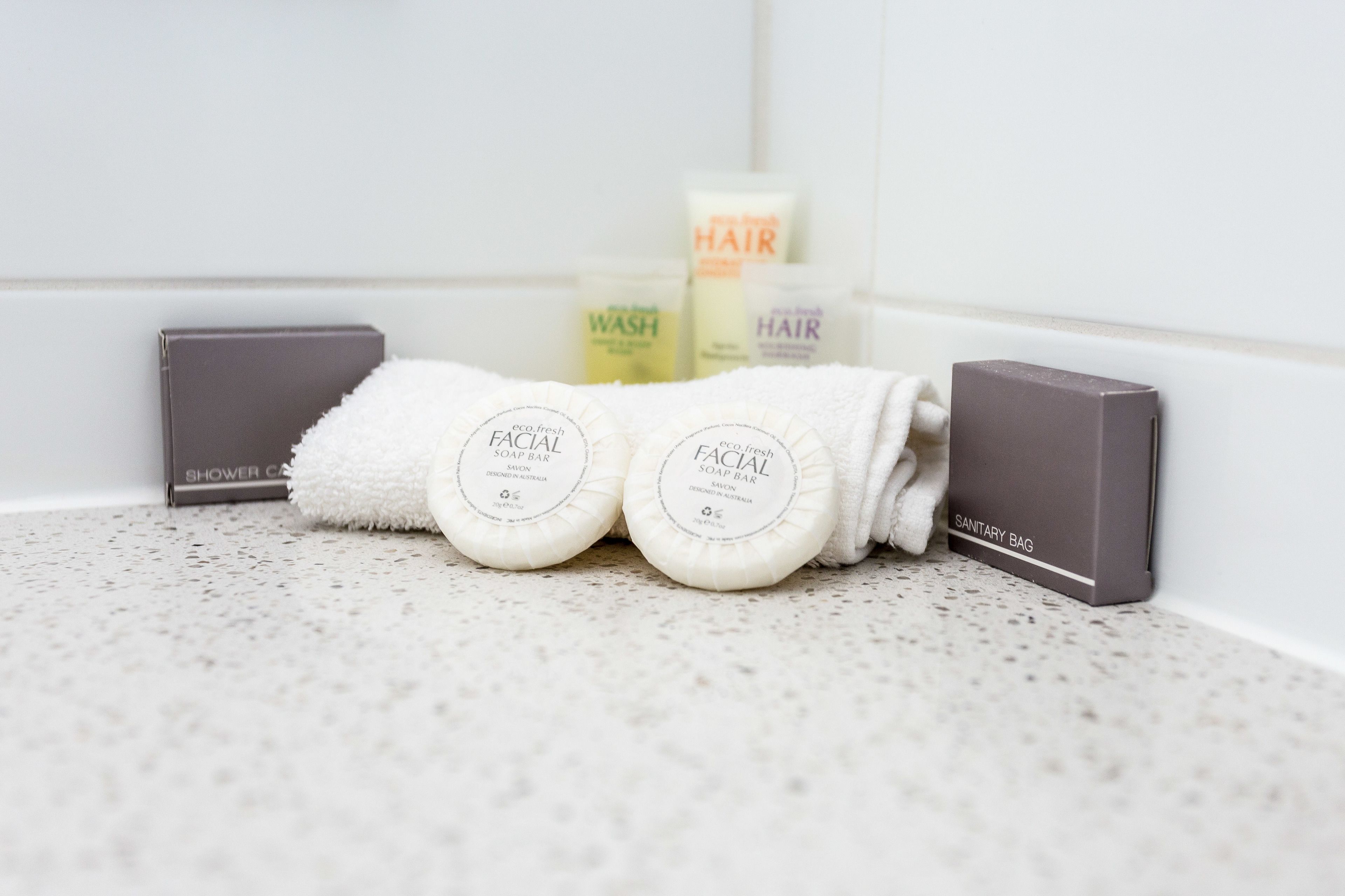 A bathroom with a white tiled wall, a shower caddy, two facial soap bars, a hair wash, and a sanitary bag.