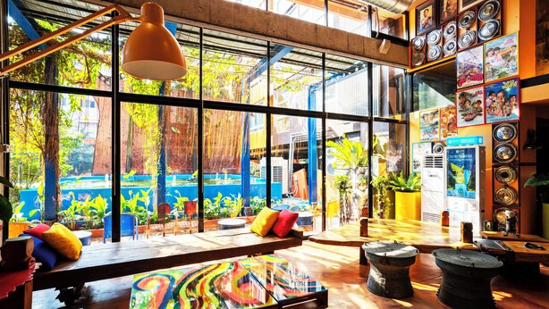 A spacious room with large windows, a wooden table, colorful artwork, and a variety of potted plants. - MeStyle Place Hotel (Bangkok)
