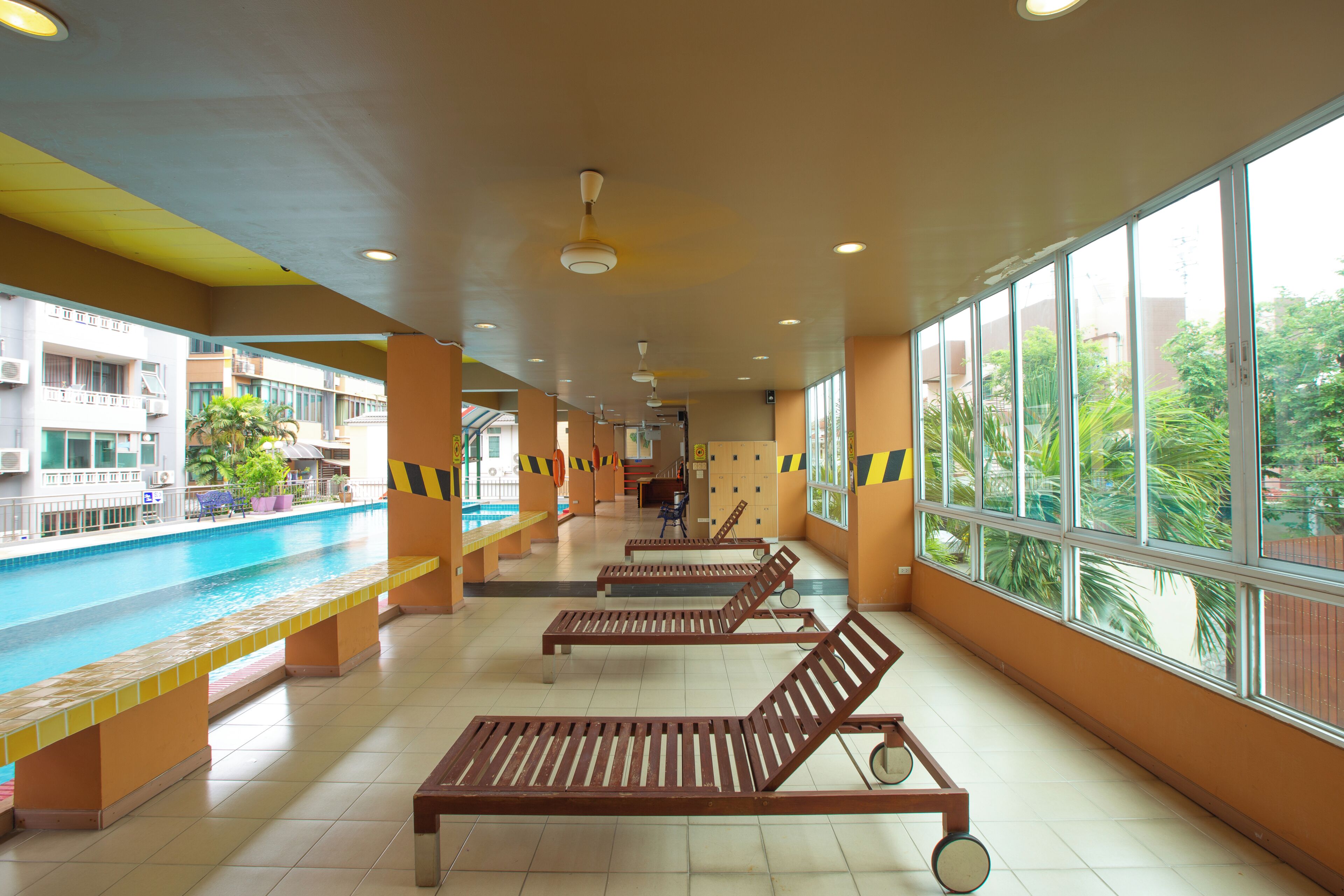 A swimming pool area with sun loungers, a tiled floor, and large windows with a view of greenery.