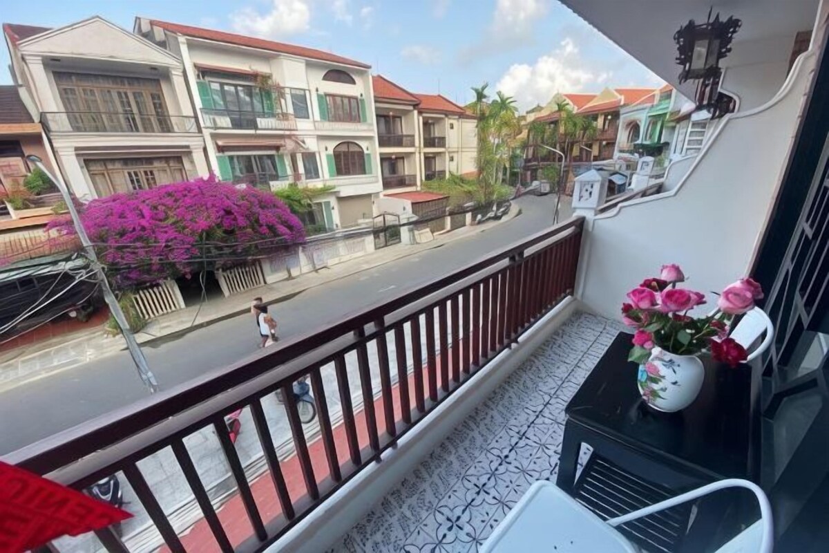 A balcony with a view of a street, a potted plant with pink flowers, and a white vase with flowers.