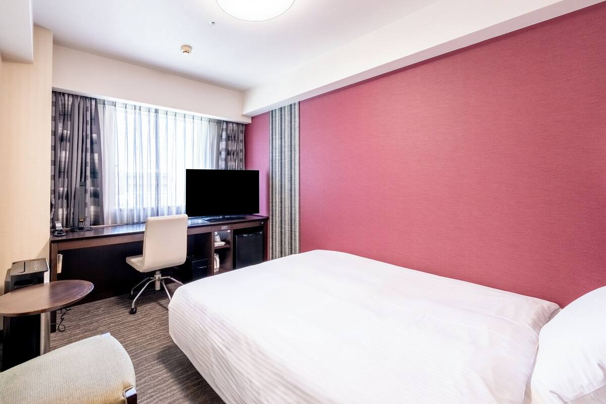 A hotel room with a bed, a desk, a chair, a TV, and a window with curtains.