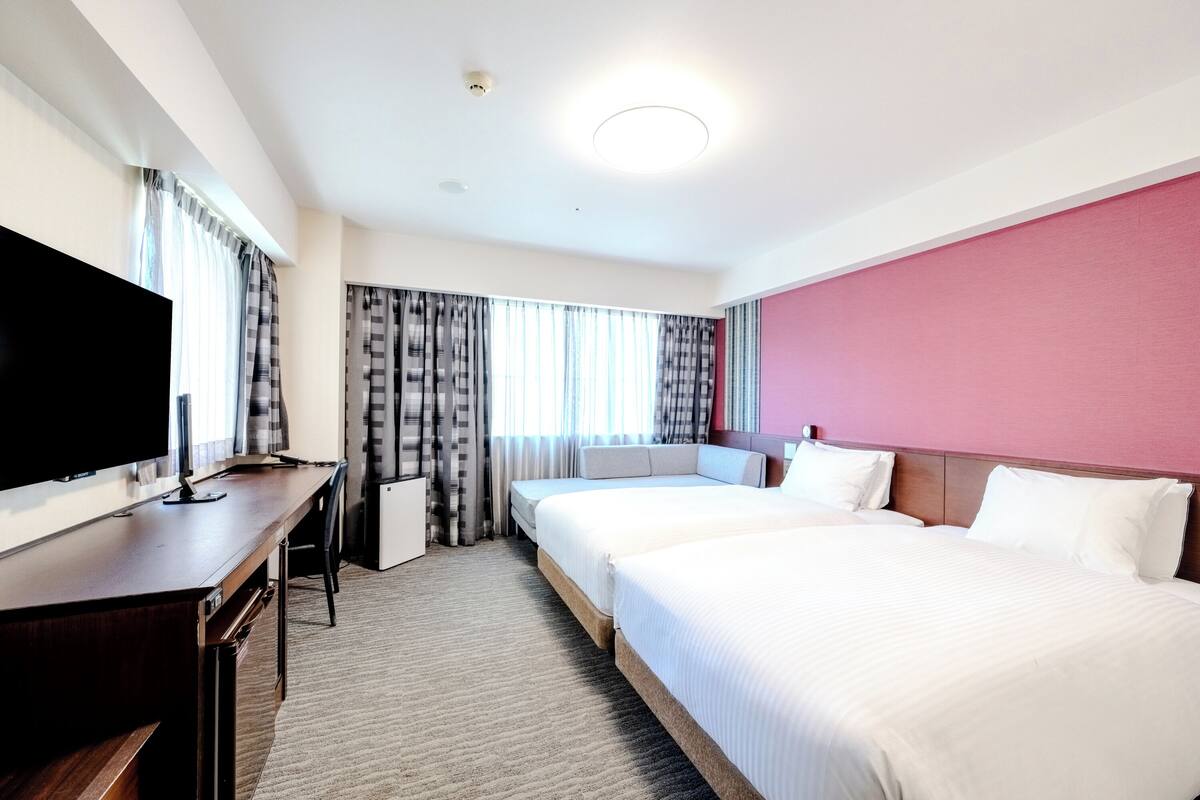 A hotel room with two beds, a desk, a flat-screen TV, and a large window with curtains.