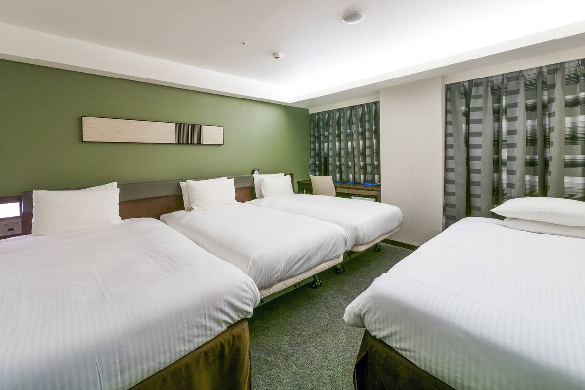 A hotel room with three beds, a green wall, a window with curtains, and a flat-screen TV.