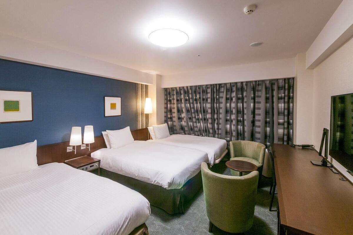 A hotel room with two beds, a desk, a chair, a television, and a large window with curtains.