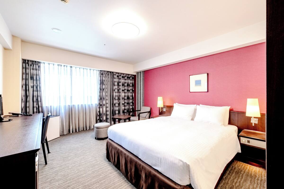 A hotel room with a bed, desk, chair, lamp, and curtains.