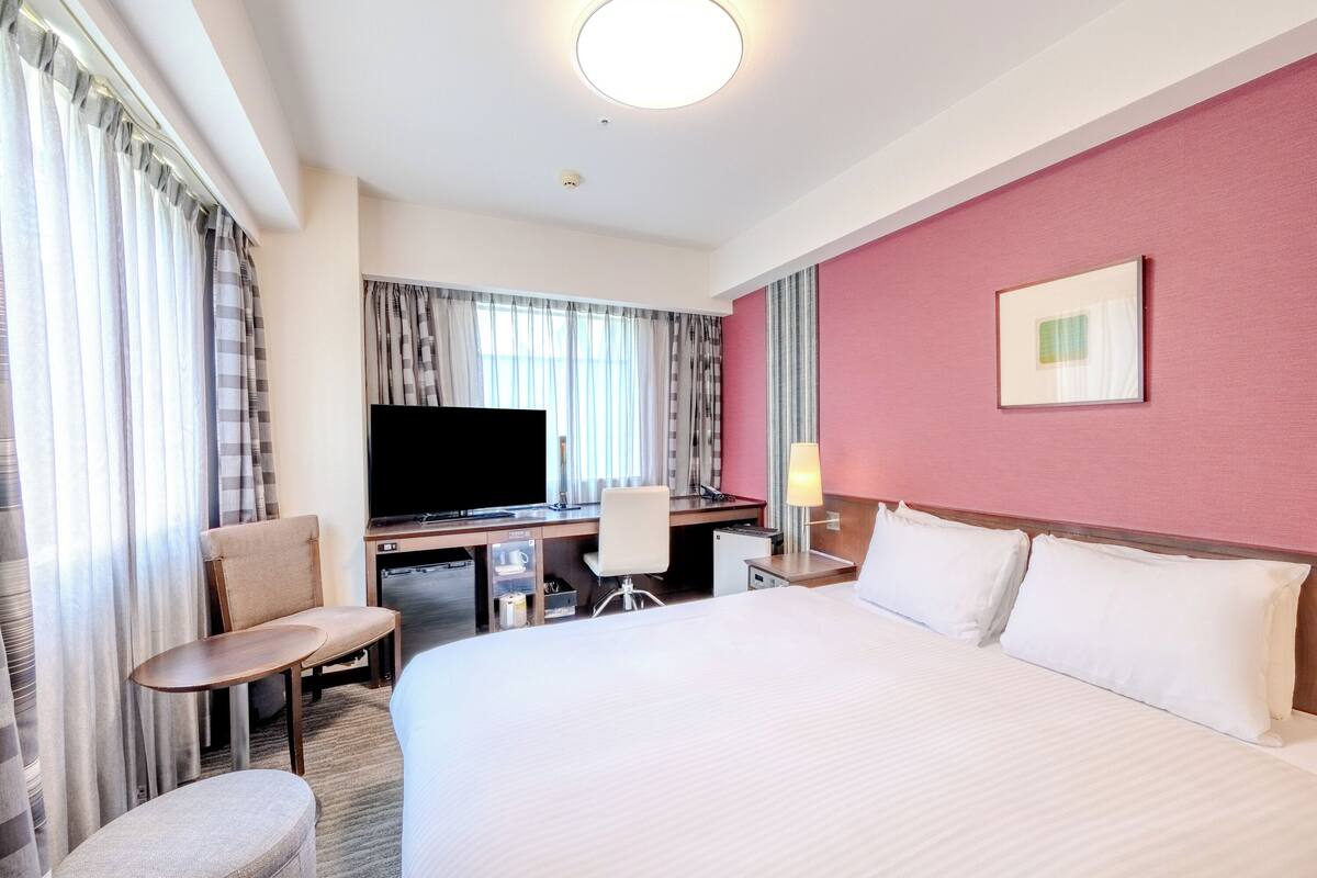 A hotel room with a bed, a desk, a chair, a television, and a lamp.