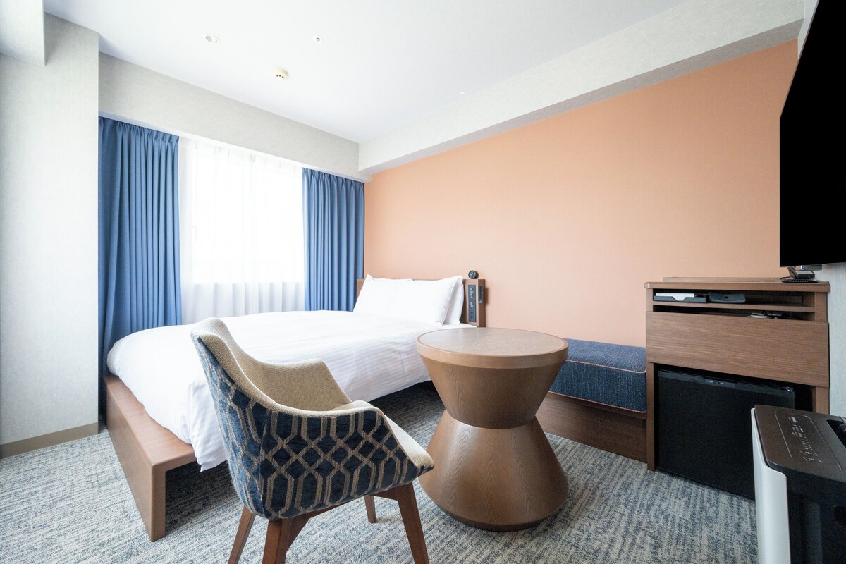 Habitación de hotel con cama, una silla, una mesita, televisión y escritorio con computadora.