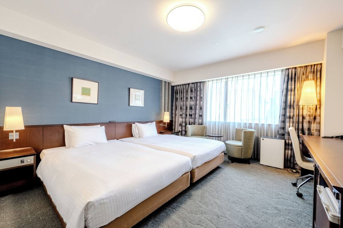 A hotel room with two beds, a desk, a chair, a lamp, a small table, and a window with curtains.