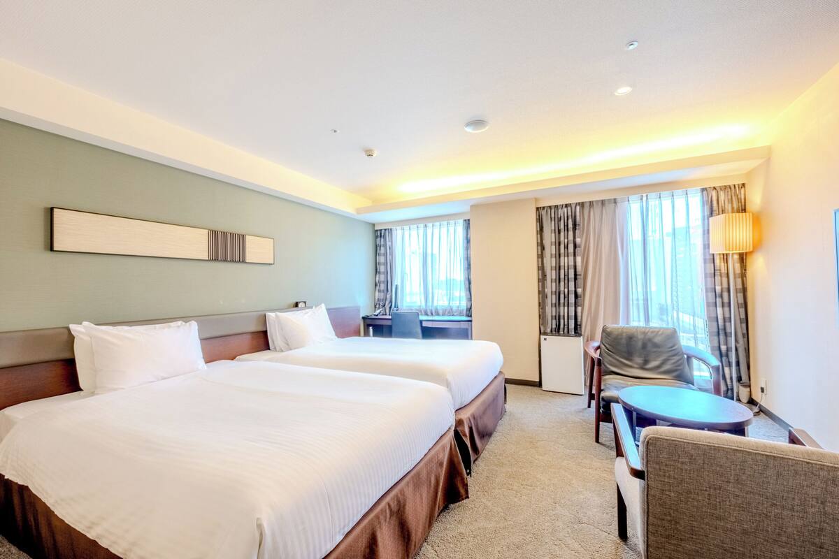 A hotel room with two beds, a desk, a chair, and a lamp.
