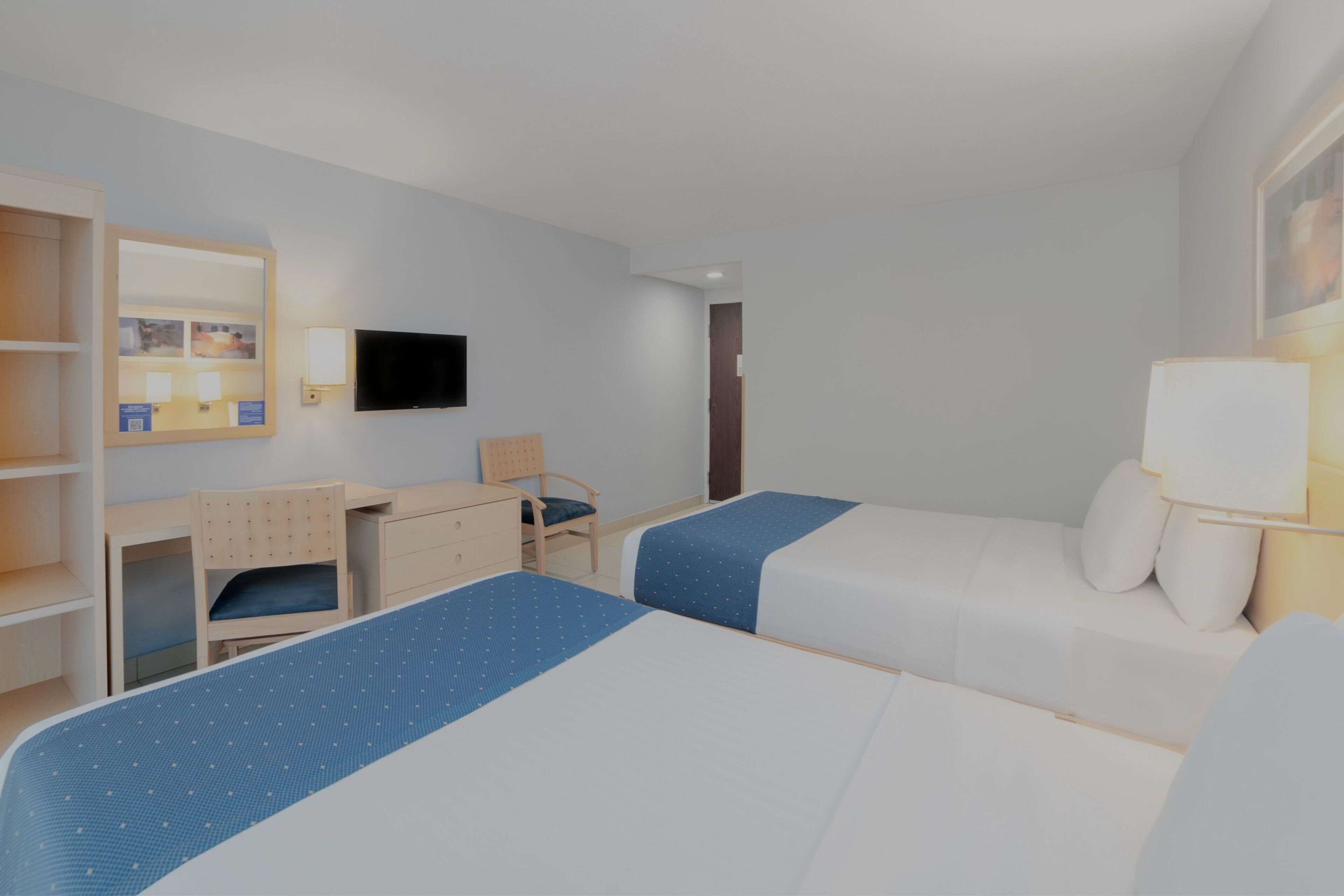 Habitación de hotel con dos camas, un escritorio, una silla, un televisor y una lámpara.