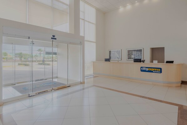 A hotel reception area with a glass door entrance, a reception desk, and a large window providing a view of the outside.