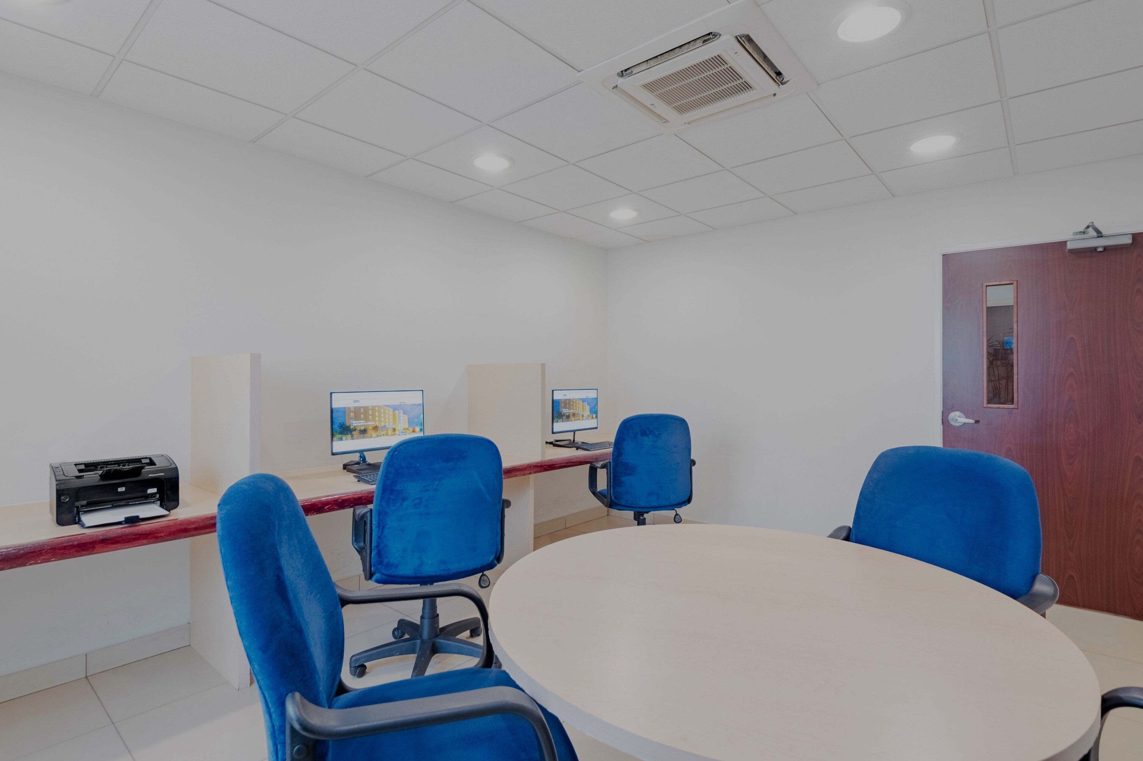 A conference room with a round table, four blue chairs, two computer monitors, and a printer on a desk.