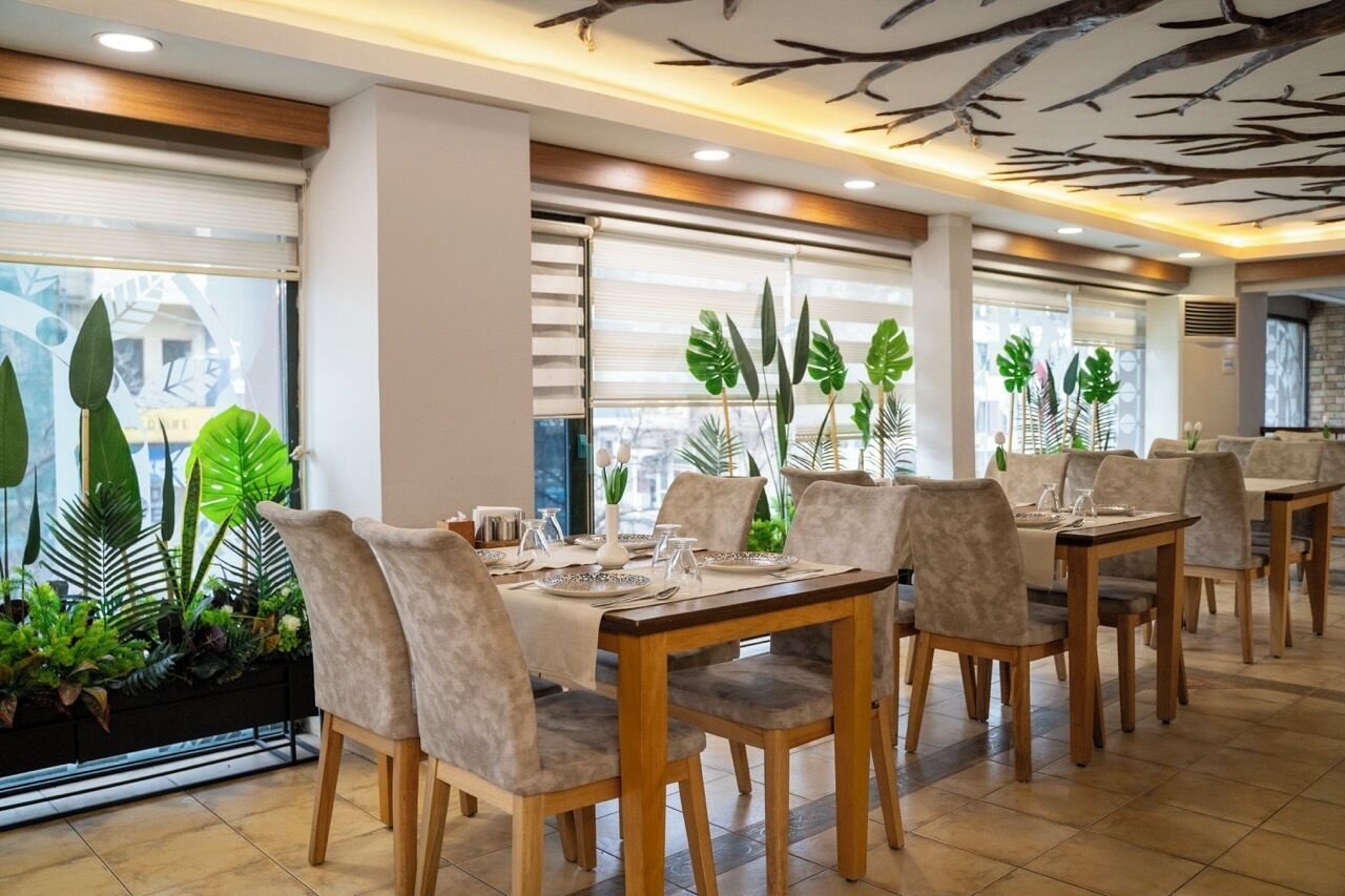 Un restaurante con varias mesas y sillas, amplias ventanas con vistas y plantas de interior.