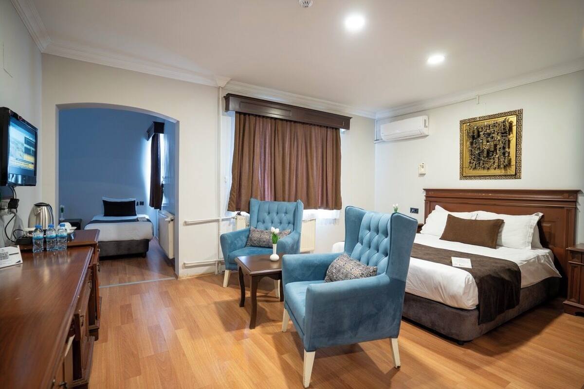 A hotel room with a bed, two blue armchairs, a wooden desk, a television, and a large decorative wall piece.